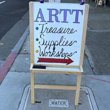 Sidewalk sign advertising art treasures, supplies, and workshops with an arrow.