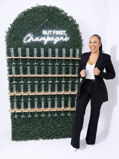 Woman in black suit poses by a champagne glass wall with neon sign.