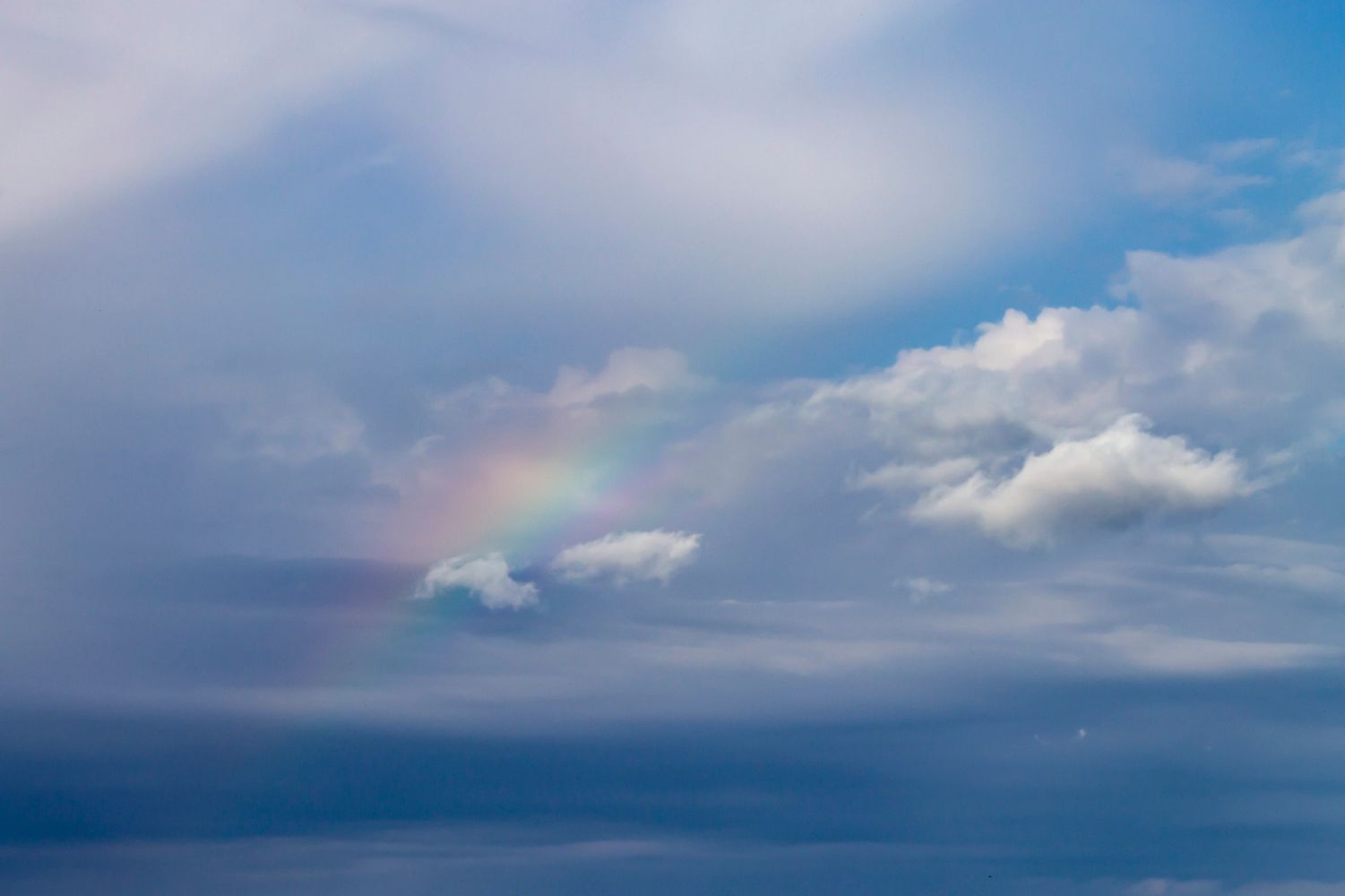 A blue sky with clouds. A rainbow appears through the clouds