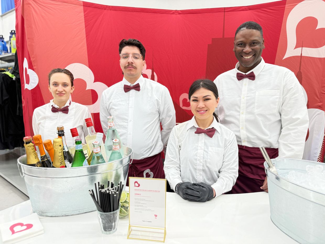 Four bartenders in white shirts and burgundy bow ties at a drink station with bottles and ice buckets.