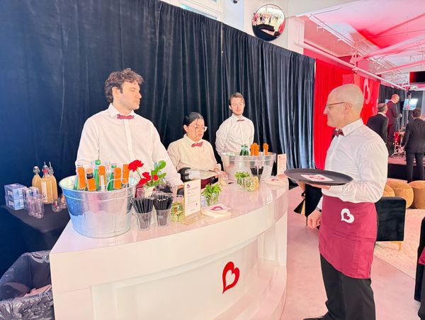 Bartenders in white shirts and red bow ties serving drinks at a sleek bar with heart logos.