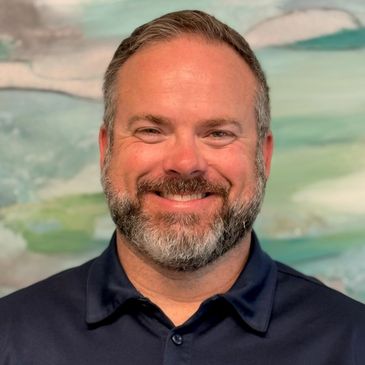 Smiling man with a beard in a navy blue shirt against a soft pastel background.