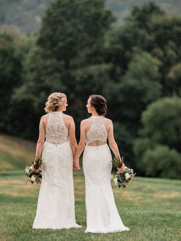 Two brides in lace wedding dresses holding hands outdoors.