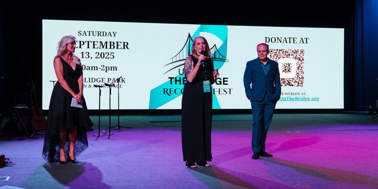 Three people on stage at a recovery fest event with a donation QR code behind.