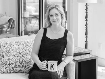 Woman in a black dress holding a mug with the letter K, sitting on a couch.