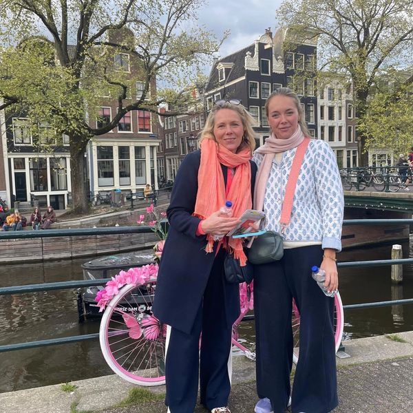 Two women posing by a canal with a decorated pink bike in a European city.