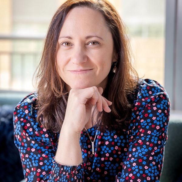 Woman with brown hair in a floral blouse, smiling thoughtfully indoors.