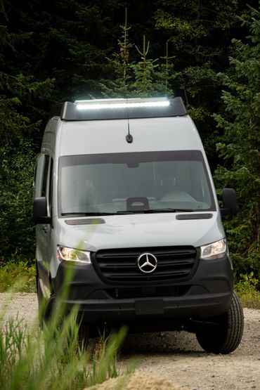 Front view of a white Mercedes van with roof light on a forest path.