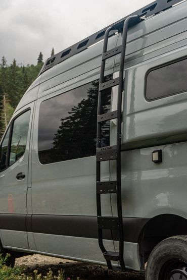A gray van with a black ladder attached to its side under a cloudy sky.