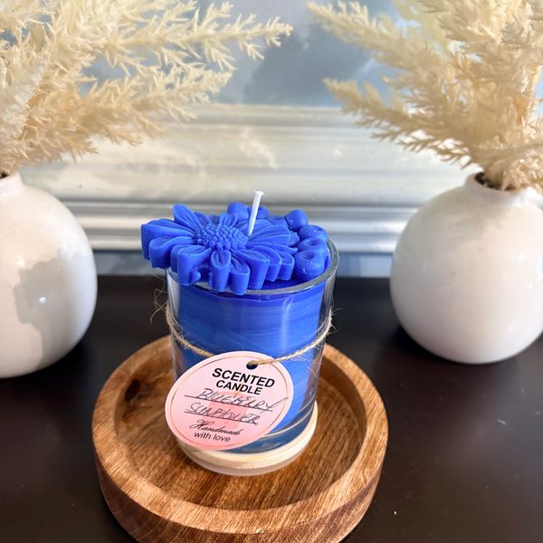 Blue floral scented candle on wooden base with white vases and dried foliage.