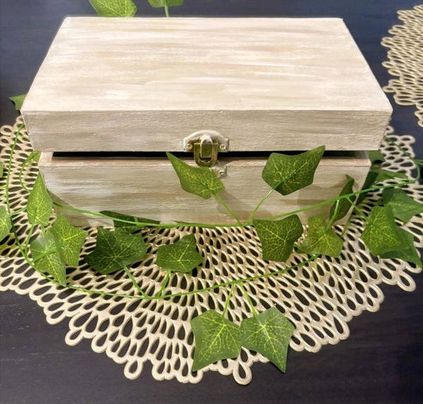 White wooden box with a latch on a decorative mat, surrounded by green leaves.
