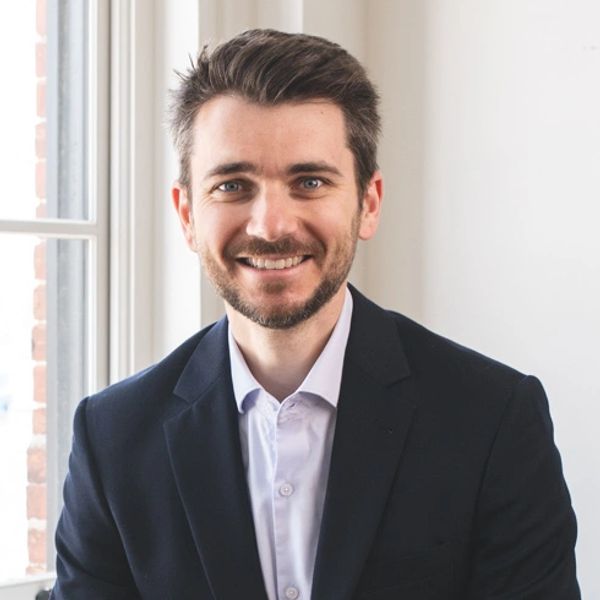 Smiling man in a suit jacket and white shirt near a window.