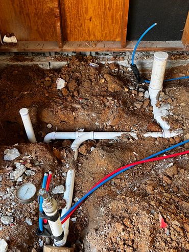 Exposed plumbing pipes and colored water lines in a dirt trench under a construction site.