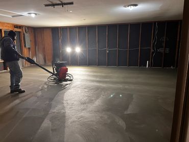 Worker smoothing a freshly poured concrete floor in a dimly lit room.