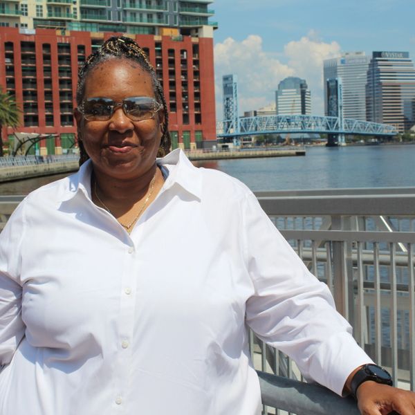 Woman in sunglasses and white shirt standing by riverside railing in urban setting.