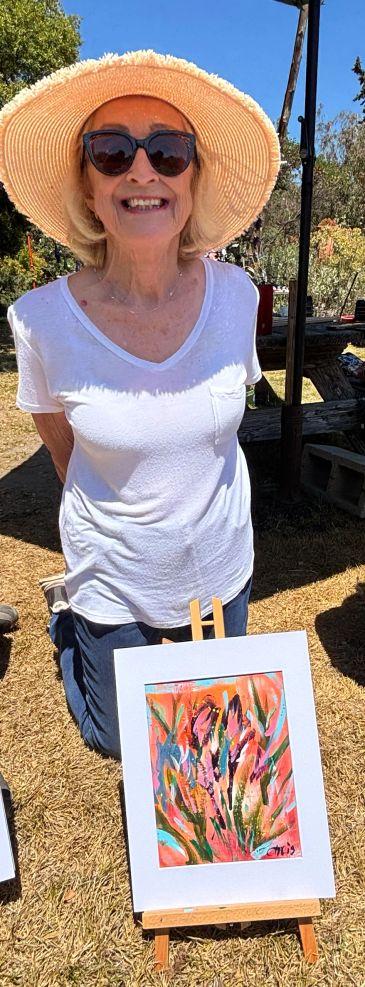 Smiling woman with a sunhat and sunglasses kneeling behind a colorful painting outdoors.