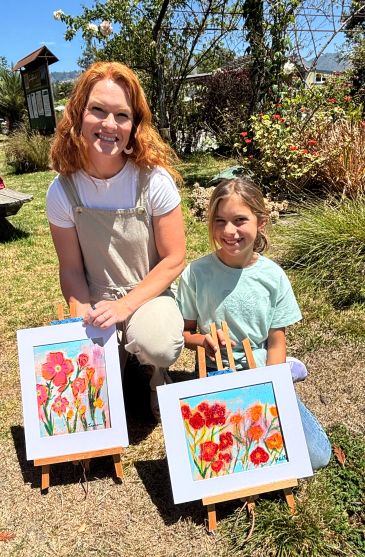 Two females outdoors showing colorful flower paintings.