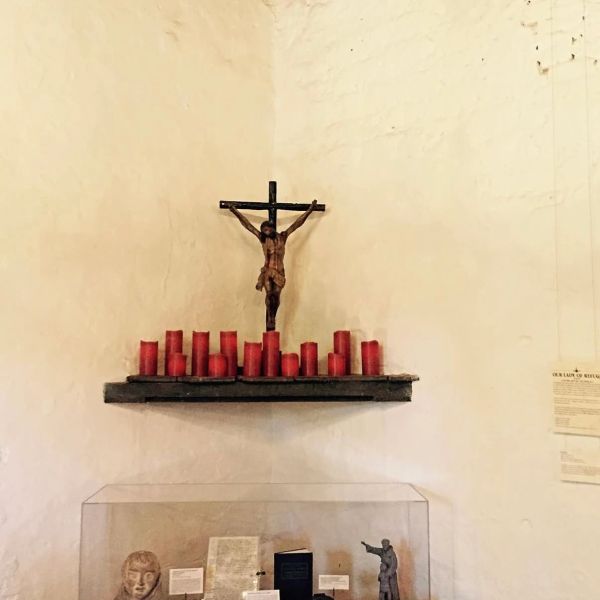 Crucifix with red candles on a shelf against a white wall.