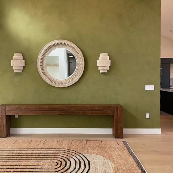 Minimalist entryway with olive green wall, wooden bench, round mirror, and layered sconces.