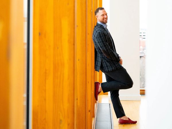 Man in checkered blazer leaning against wooden wall, smiling.