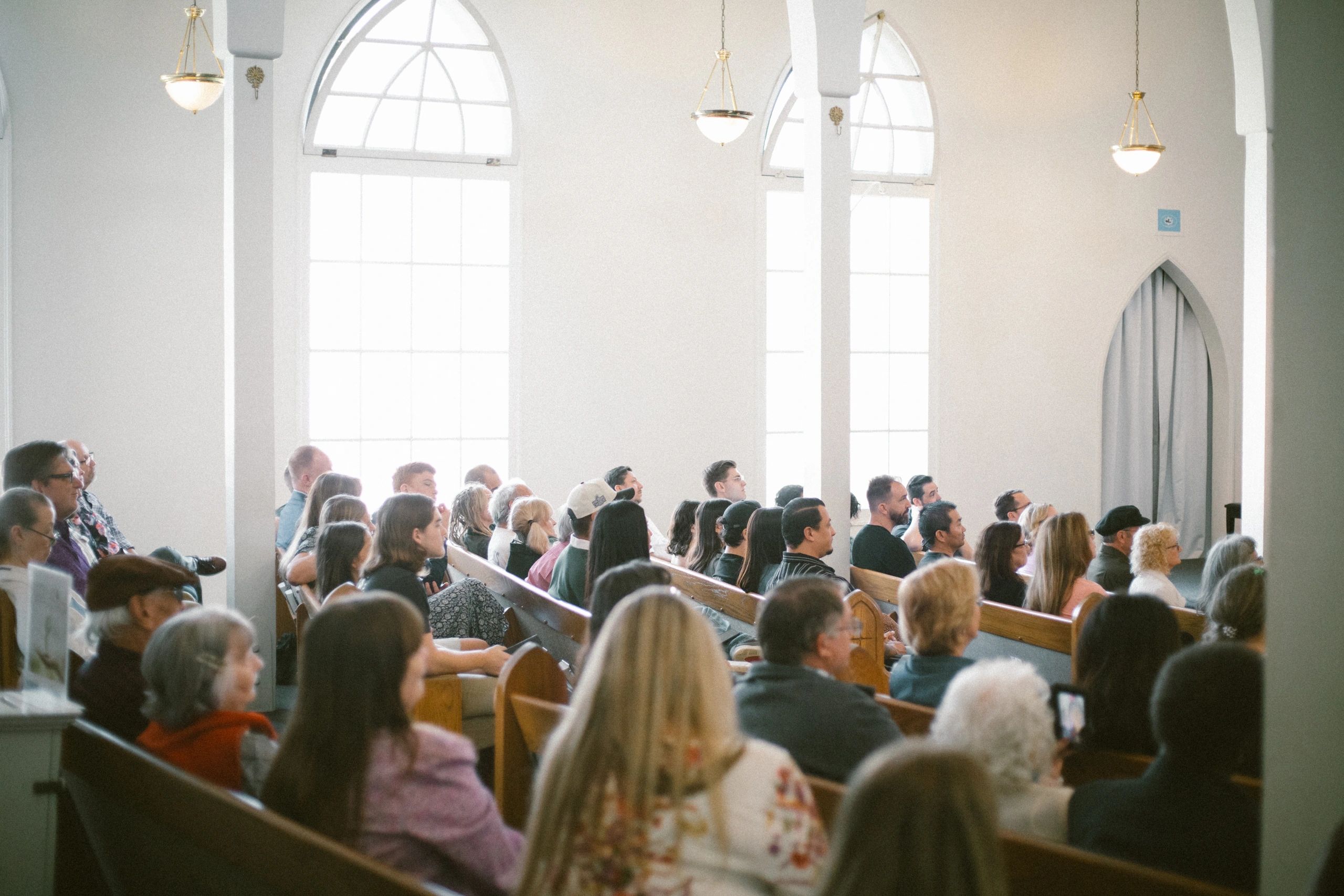 People attentively listening inside a bright church with arched windows.