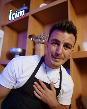 Man in apron smiling with hand on chest in a kitchen.