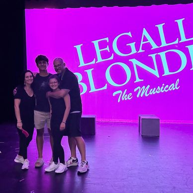 A group of four people posing in front of a bright pink 'Legally Blonde The Musical' backdrop.