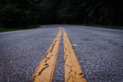 Image of cracked blacktop road with double yellow lines in greed wooded area.