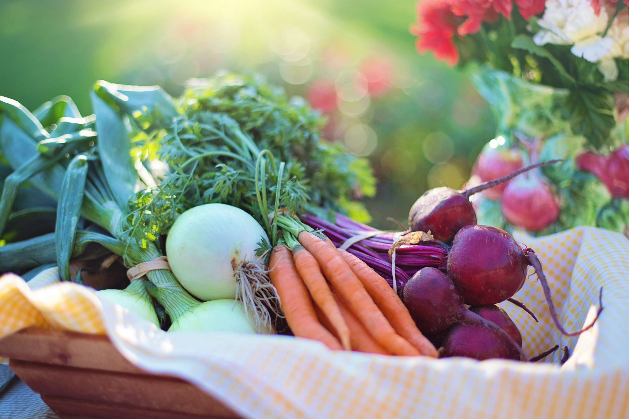 Onions, carrots, and radishes in a wood square basket with yellow and white checkered linen cloth.