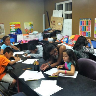 Teacher assisting a young girl with her homework in a busy classroom.