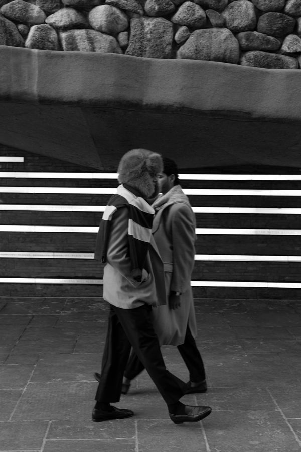 Two people walking past a stone wall and striped background in black and white.