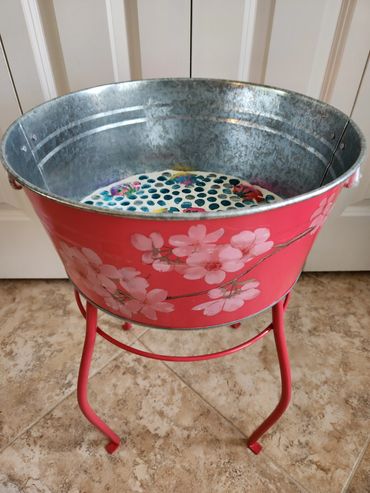 Red metal tub with pink flower design on a matching stand indoors.