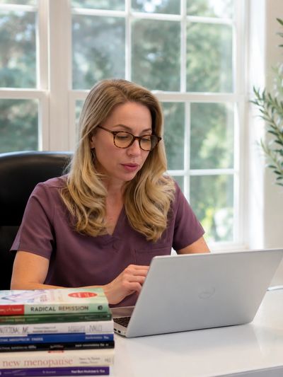 Woman in glasses working on a laptop at a desk with a stack of health books.