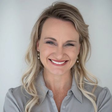Smiling woman with blonde hair wearing pearl earrings and a gray blouse.