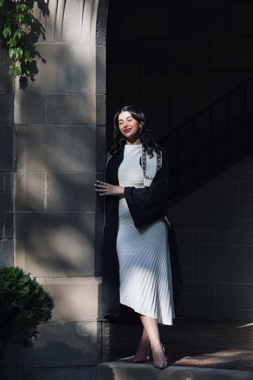 A woman in a white dress and black graduation gown stands by a stone archway in sunlight.