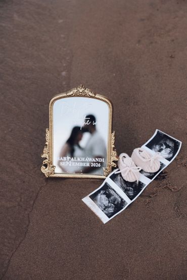 Mirror reflecting a couple with baby announcement and ultrasound photos on sand.