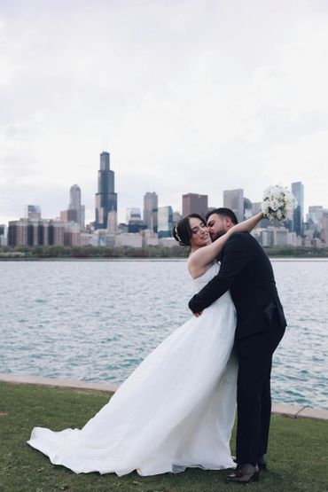 Bride and groom embracing by the water with city skyline backdrop.