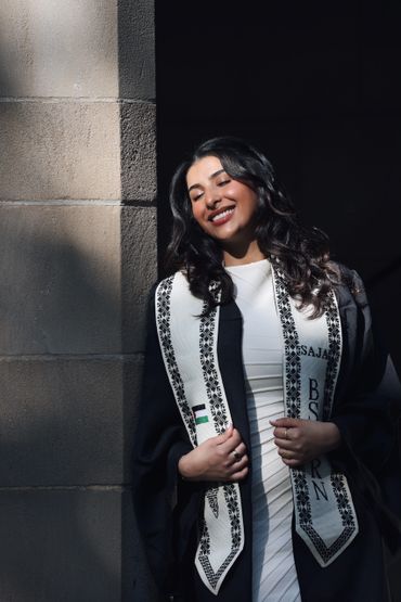 Woman in graduation attire smiling in sunlight near a stone wall.