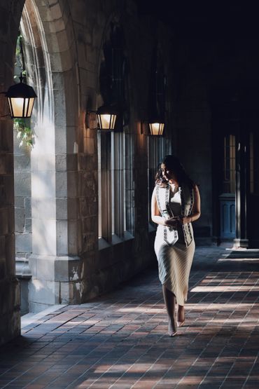 A woman walks through a sunlit stone corridor with arched windows.