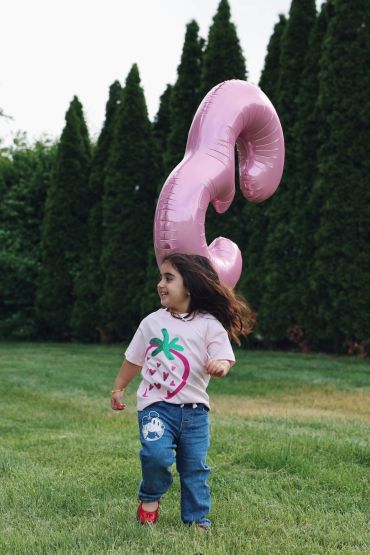 A young girl running outdoors with a large pink balloon shaped like the number 3.