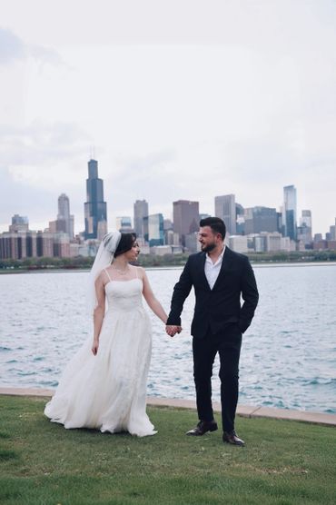 Bride and groom holding hands by the water with a city skyline behind them.