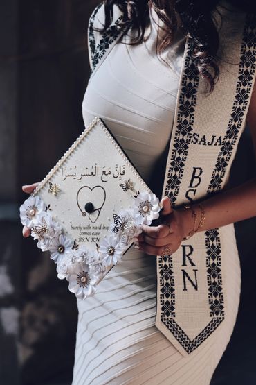 A graduate in a white dress holding a decorated cap and a BSN RN sash.