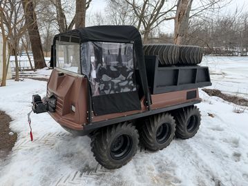 An amphibious six-wheeled vehicle parked on snow near bare trees.
