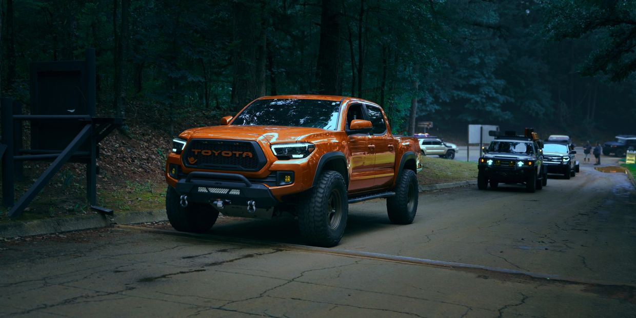 Orange Toyota truck parked on a forest road with other vehicles behind it.