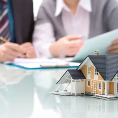 A miniature house model on a table with two people discussing documents in the background.