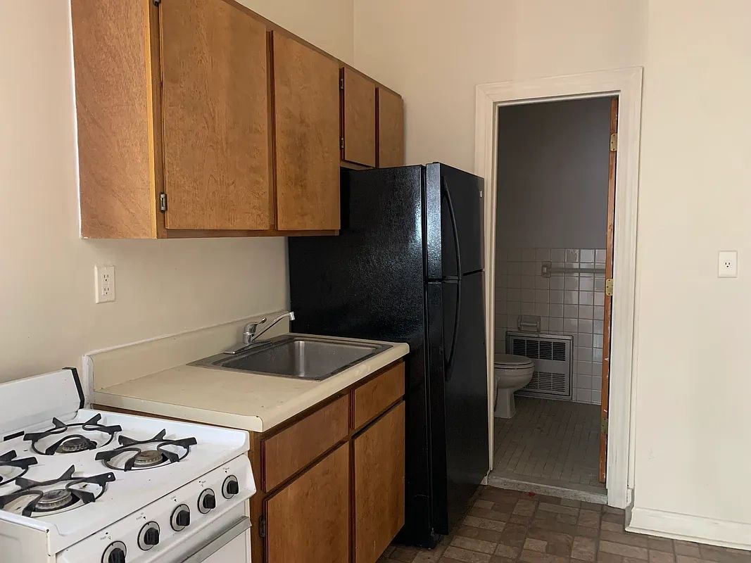 Small kitchen with wooden cabinets, white stove, black refrigerator, and a sink next to a bathroom door.