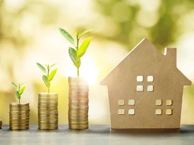 Stacks of coins with growing plants beside a house model symbolizing financial growth and investment.