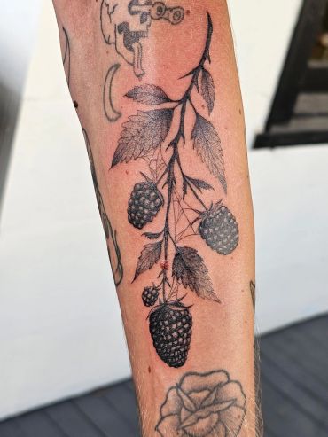 Detailed tattoo of a blackberry branch with leaves on a forearm.