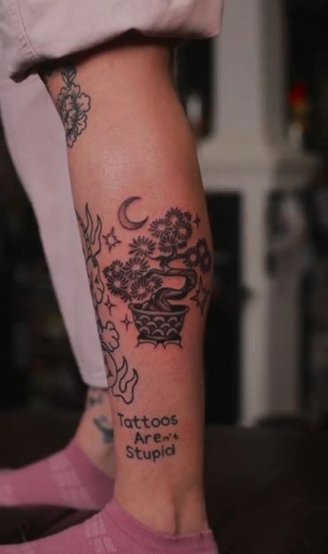 Black ink tattoo of a bonsai tree with stars and moon, and text 'Tattoos Aren't Stupid' on a leg.