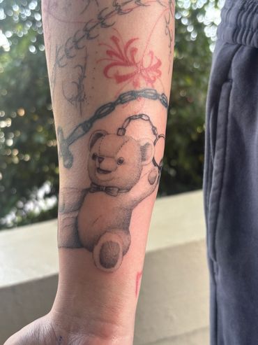 A realistic teddy bear tattoo on a forearm with other designs around it.